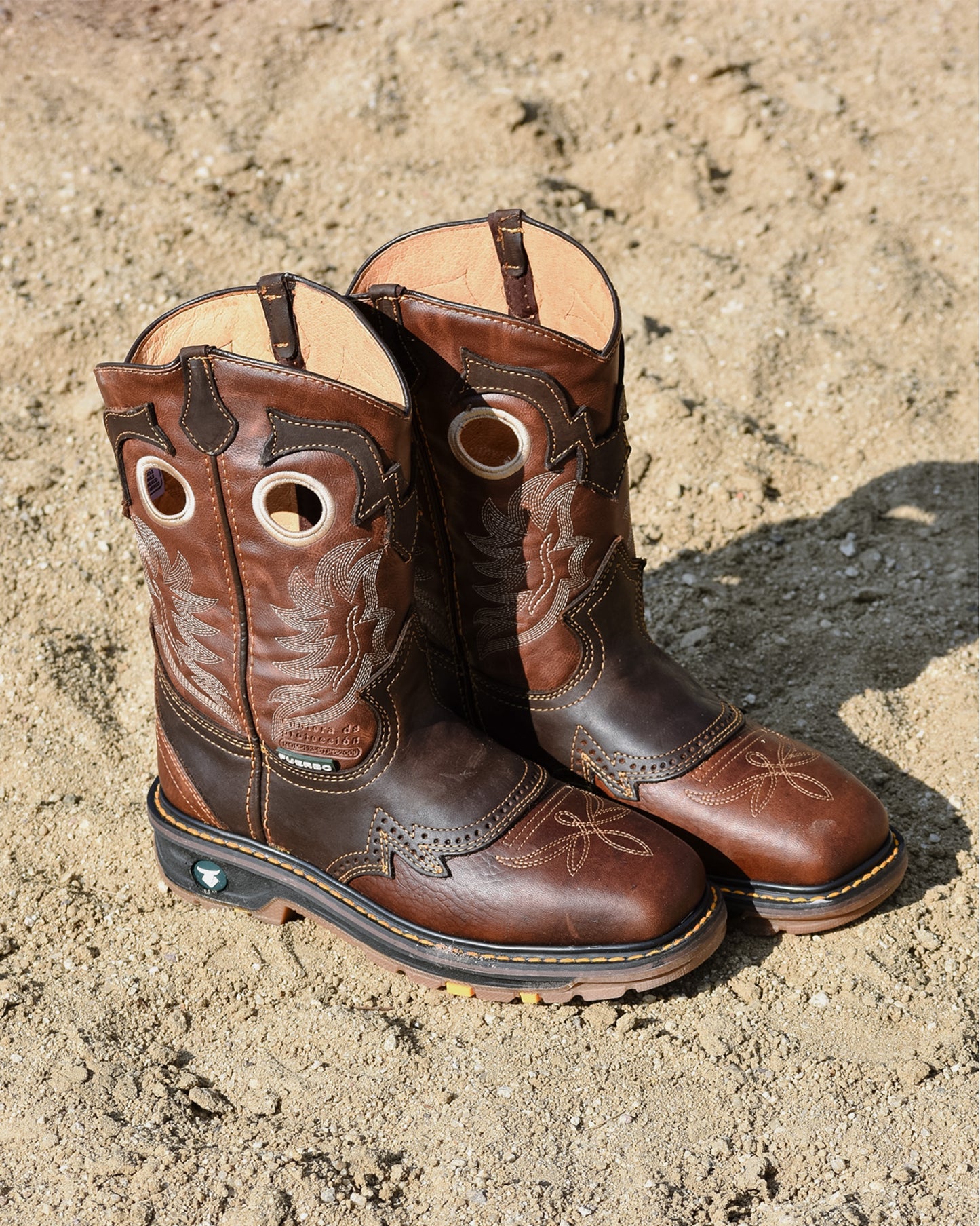 A pair of brown leather western boots with decorative stitching and circular cutouts on the shaft, photographed standing on sandy ground outdoors.